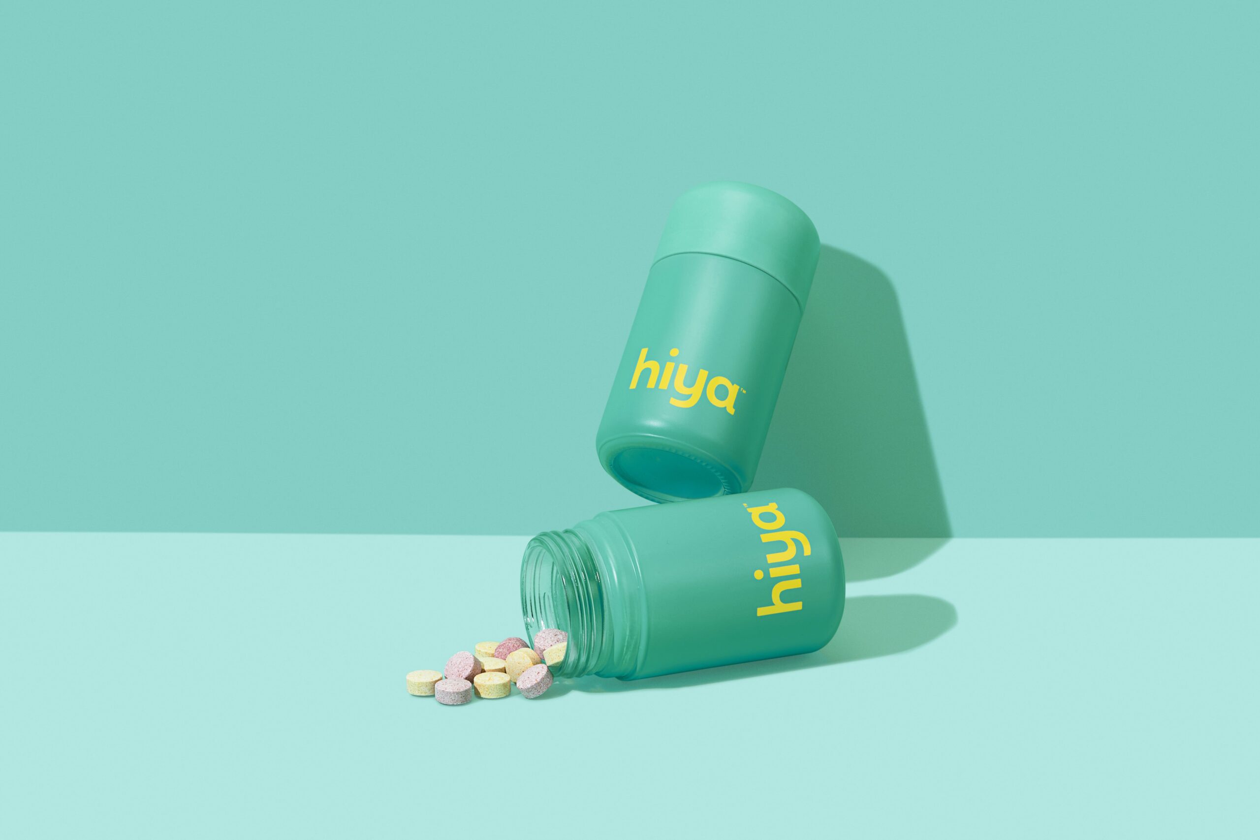 A bottle of Hiya's Daily Probiotic, tipped over with several multicolored chewable tablets spilling out onto a similarly colored turquoise background.