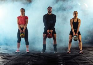 Three athletes in a smoke-filled gym holding kettlebells, wearing heat-trapping workout gear.