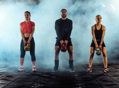 Three athletes in a smoke-filled gym holding kettlebells, wearing heat-trapping workout gear.
