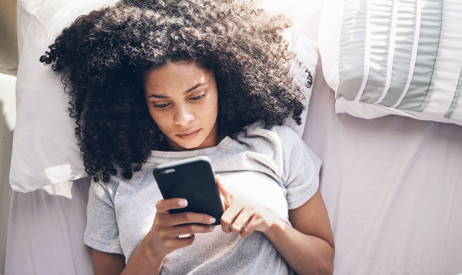 A woman with curly hair lying on a bed and looking at her phone.
