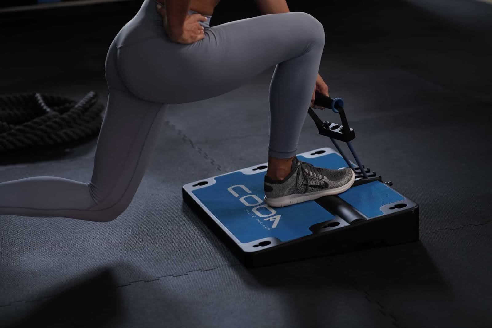 A close-up of a person’s legs in gray leggings using the COBA Board Plus™. The person is performing an exercise with one knee on the board and the other leg extended back.