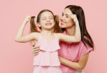 A smiling mother embraces her daughter, who playfully flexes her arms to show off her strength.