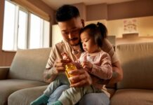 Father and young daughter sitting on a couch, holding a vitamin bottle together as the child reaches for a kids multivitamin.
